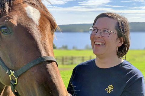 Kvinna står i t-shirt med Wångens emblem på utomhus en solig dag. Hon ler och tittar på en varmblodstravare i grimma. I bakgrunden syns gröna hagar och en sjö.