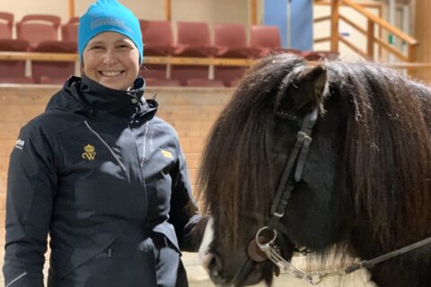 Kvinna i turkos mössa och blå jacka ler mot kameran. Hon står i ett ridhus bredvid en svart islandshäst